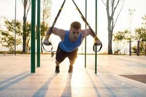 Training mit Aufhängegurten im Fitnessstudio im Freien, starkes Manntraining am frühen Morgen im Park, Sonnenaufgang oder Sonnenuntergang im Meereshintergrund foto