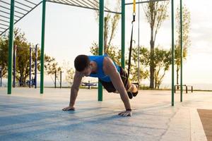 Training mit Aufhängegurten im Fitnessstudio im Freien, starkes Manntraining am frühen Morgen im Park, Sonnenaufgang oder Sonnenuntergang im Meereshintergrund foto