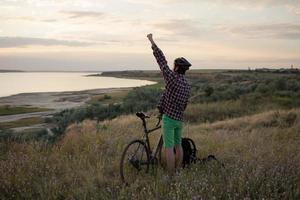 Silhouette eines Mannes mit Touring-Rennrad beobachten und Foto des Sonnenuntergangs im See auf dem Handy machen