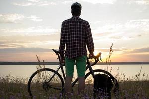 Silhouette eines Mannes mit Touring-Rennrad beobachten und Foto des Sonnenuntergangs im See auf dem Handy machen