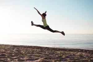 junge professionelle turnerin tanzt am strand, trainingsübungen mit coolen junps, sonnenaufgang im meer- oder ozeanhintergrund foto
