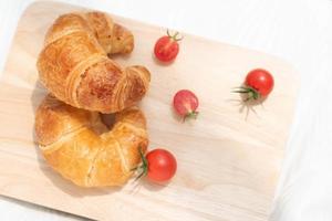 Croissant mit roten Tomaten isoliert auf Holzplatte, frisch gebackenes Brot für ein einfaches Frühstück. foto