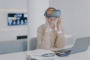 Aufgeregtes Mädchen im vr-Headset, das am Schreibtisch vor dem Laptop sitzt. Arbeiten an einem Projekt im Cyberspace. foto