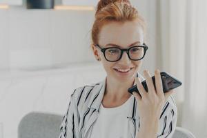 Schöne positive rothaarige Frau mit Brille, die ein Mobiltelefon hält und eine Audionachricht sendet foto