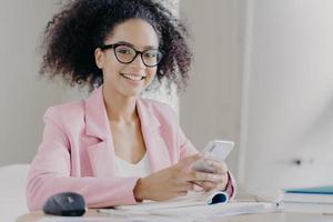 positive lockige frau trägt eine brille, formelle kleidung, hält ein modernes handy in der hand, sucht informationen über geschäftsnachrichten, sitzt an schreibtischpapieren, trinkt kaffee, hat einen glücklichen ausdruck, lächelt foto