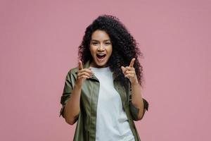 Fröhliche, selbstbewusste Frau mit Afro-Haaren zeigt mit dem Zeigefinger direkt auf die Kamera, wählt Sie aus, wählt jemanden in ihrem Team aus, gekleidet in weißes T-Shirt und Lederhemd, posiert vor rosafarbenem Hintergrund foto