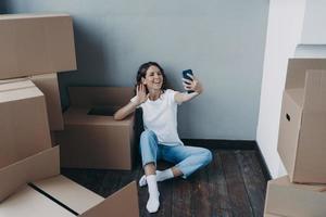 Fröhliche junge Spanierin in Jeans und weißem T-Shirt, die Kisten auspackt und einen Videoanruf am Telefon hat. foto