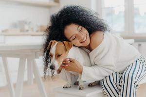 schöne junge lockige afroamerikanerin umarmt geliebten rassehund mit liebe, hat ein sanftes lächeln, trägt stilvolle kleidung, posiert vor häuslichem hintergrund in einer modernen wohnung, drückt fürsorge aus foto