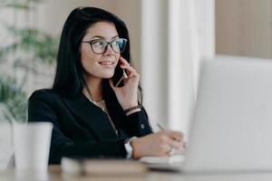 Elegante Unternehmerin mit zufriedenem Gesichtsausdruck führt ein Telefongespräch, überprüft die Agenda, während sie an einem modernen Laptop arbeitet, schreibt Notizen in den Notizblock, posiert im Coworking Space foto