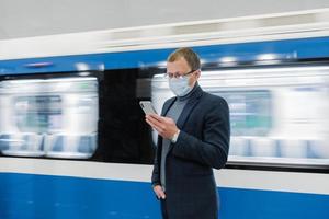 junger mann manager benutzt handy, verhindert die ausbreitung von coronavirus, posiert gegen u-bahn, posiert am bahnsteig, überprüft nachrichten online, trägt medizinische maske. epidemische situation, gesundheitskonzept foto