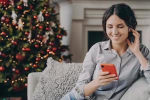 Entspannte, glückliche italienische Frau, die während der Weihnachtsferien zu Hause auf einem gemütlichen Sofa mit Telefon sitzt foto