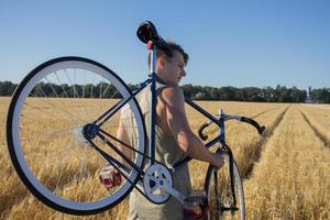 Junger Mann fährt Fahrrad mit festem Gang auf der Landstraße, den Feldern und dem blauen Himmelshintergrund foto