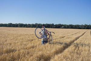 Junger Mann fährt Fahrrad mit festem Gang auf der Landstraße, den Feldern und dem blauen Himmelshintergrund foto