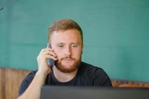 Junge bärtige Rothaarige arbeiten im Café mit Handy und Laptop foto