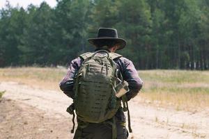 Rückansicht eines jungen männlichen Wanderers mit Cowboyhut, der im Sommertag im Freien spazieren geht. Hipster-Reisender. foto