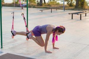 schöne fitte frau in rosa und lila sportbekleidung morgens training im outdoor-fitnessstudio, übungen mit aufhängungsgurten im park foto