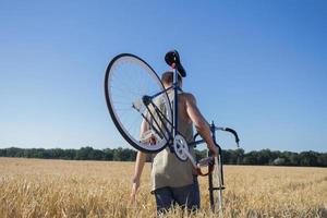 Junger Mann fährt Fahrrad mit festem Gang auf der Landstraße, den Feldern und dem blauen Himmelshintergrund foto