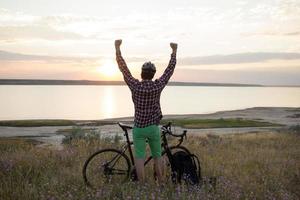 Silhouette eines Mannes mit Touring-Rennrad beobachten und Foto des Sonnenuntergangs im See auf dem Handy machen