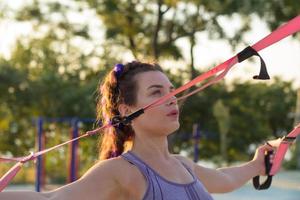 schöne fitte frau in rosa und lila sportbekleidung morgens training im outdoor-fitnessstudio, übungen mit aufhängungsgurten im park foto