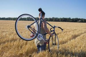 Junger Mann fährt Fahrrad mit festem Gang auf der Landstraße, den Feldern und dem blauen Himmelshintergrund foto