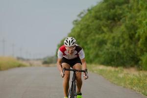 Radrennfahrer im Helm- und Sportbekleidungstraining allein auf leerer Landstraße, Feldern und Bäumen im Hintergrund foto