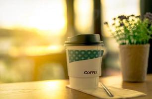 Kaffeeglas aus Papier im Café auf verschwommenen Blumen mit grünen Blättern im Topf auf braunem Holztisch im Café am Morgen mit Sonnenlicht. Einweg-Pappbecher. grüne Verpackung. heisses Getränk.