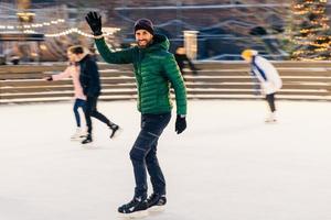 glücklicher mann winkt mit der hand, als sein freund auf der eisbahn bemerkt, hat lust zusammen skaten zu gehen, freut sich über das treffen, hat gute zeit und stimmung, unterhält sich. Menschen und Winteraktivitäten foto