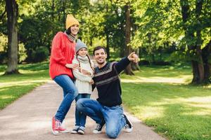 Horizontales Porträt einer Familie mit guter Beziehung stehen nahe beieinander, bewundern Landschaften oder wunderschöne Natur. Der glückliche Vater zeigt seiner Tochter einen großen alten Baum im Park, zeigt mit dem Zeigefinger an foto