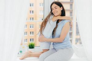 menschen, beziehung und elternschaftskonzept. positive liebevolle Frau mit angenehmem Aussehen, trägt lässige Haushaltskleidung, umarmt ihre kleine Tochter, drückt Liebe aus, erzieht ihr Kind foto