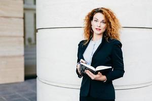 porträt einer geschäftsfrau mit lockigem haar, rot bemalten lippen, eleganter kleidung, die in ihr tagebuch schreibt. gutaussehendes damenmodell mit attraktivem aussehen, das ihr notizbuch mit stift hält foto