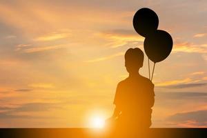 Silhouette eines Jungen, der allein sitzt und den Sonnenuntergang beobachtet und einen Ballon in der Hand hält. foto