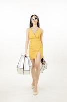 Selbstbewusste asiatische Frau in einem schwülen gelben Kleid, das eine Sonnenbrille trägt und eine Einkaufstasche trägt, hat Spaß beim Einkaufen. foto