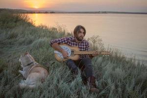 Folk- oder Country-Gitarrist im Wald mit Hund, Mann im Wald, Lagerfeuer und Sonnenuntergang im Hintergrund foto