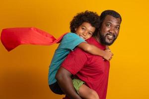 Afrikanischer Vater und Sohn spielen tagsüber Superhelden. Leute, die Spaß am gelben Hintergrund haben. Konzept der freundlichen Familie.