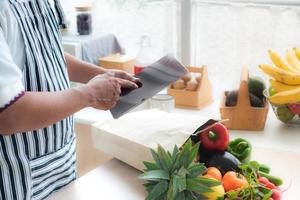 Nahaufnahme der Hand des Küchenchefs mit Tablet, um Obst und Gemüse online zu kaufen. und es gibt obst und gemüse und papiertüten auf dem küchentisch. online-einkaufs- und lieferkonzept. foto