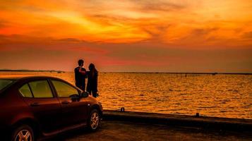 Silhouette eines Glückspaares, das bei Sonnenuntergang am Auto am Meer steht. schöner orangefarbener Himmel und Wolken foto