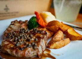 Selektiver Fokus auf Steak mit Soße und Pfeffer serviert mit gebratenen Kartoffeln und gedämpften Karotten, Blumenkohl und Brokkoli. Steak auf weißem Teller auf Holztisch gelegt. Tomahawk-Steak. foto