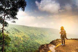 junge reisende frau mit rucksackhut und kamera in der rechten hand steht auf der spitze der bergklippe und beobachtet nach dem regen in ihrem urlaub einen schönen blick auf wald und himmel. asiatische Frau allein reisen. foto