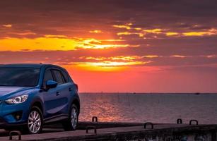 blaues kompaktes suv-auto mit sport und modernem design, das bei sonnenuntergang auf einer betonstraße auf dem meer geparkt ist. umweltfreundliche Technologie. Hybrid- und Elektroautotechnik. Pkw-Stellplatz. sommer reisen. foto