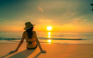 Rückansicht der asiatischen Frau trägt Bikini und Strohhut, sitzt am Sandstrand, entspannt sich und genießt den Urlaub am tropischen Paradiesstrand. Frau sieht schönen Sonnenuntergang. Sommerurlaub. alleine reisen. sommergefühl. foto