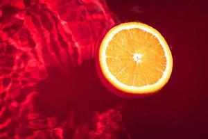 Stück frische Orange auf der Wasseroberfläche mit Wellen und Reflexionen. Schwimmen Stück Orange auf rotem abstrakten Hintergrund. foto