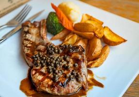 Steak mit Soße und Paprika serviert mit gebratenen Kartoffeln und gedämpften Karotten, Blumenkohl und Brokkoli. Steak auf weißem Teller mit Edelstahlgabel und Messer auf Holztisch gelegt. Tomahawk-Steak. foto