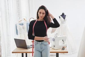 berührt Haare. Porträt einer Modedesignerin, die in der Werkstatt und am Tisch mit Instrumenten dahinter steht foto