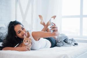 Selfies machen. attraktive Blondine, die mit ihrem süßen Kätzchen auf dem weißen Bett ruht foto