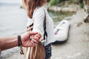 Porträt einer glücklichen jungen Frau mit Hut, die den Mann mit der Hand hält und zum Boot auf dem See in den Bergen geht foto