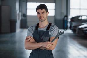 ruhig und konzentriert. Porträt eines ernsthaften Arbeiters in Uniform, der mit einem Schraubenschlüssel in der Hand in seiner Werkstatt steht foto