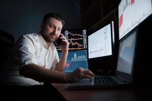 nettes Gespräch. bärtiger mann im weißen hemd hat einen geschäftsanruf und arbeitet im büro mit mehreren computerbildschirmen in indexdiagrammen foto