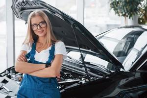 stolz auf erfolgreiche Arbeit. Die Werkstattfrau im weißen Hemd und in der blauen Uniform steht mit aufgesetzter Motorhaube vor einem schwarzen Auto foto