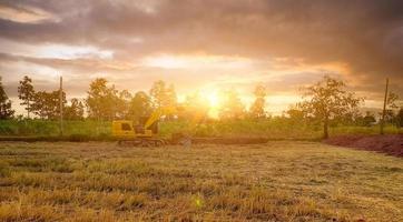 Landschaft aus Reisfeldern und tropischen Wäldern mit Sonnenuntergangshimmel. Baggerarbeiten durch Graben von Erde. Bagger, der nach der Ernte im Winter auf dem Reisfeld auf dem Boden gräbt. Aushubmaschine. Aushubfahrzeug. foto