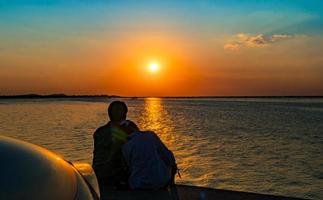 Silhouette des Glückspaares, das am Strand vor dem Auto mit orangefarbenem und blauem Himmel bei Sonnenuntergang sitzt und sich entspannt. sommerferien und reisekonzept. romantisches junges Paar aus am Meer.